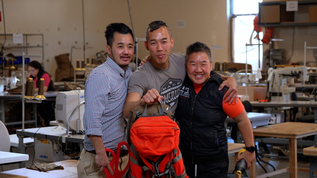 Three people are standing in a room, posing with a backpack.